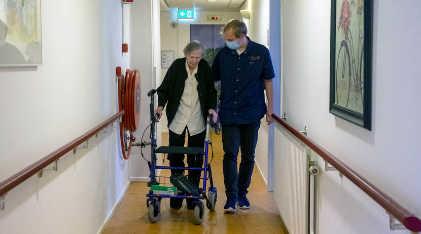 Bewoner met rollator en medewerker lopen samen op de gang